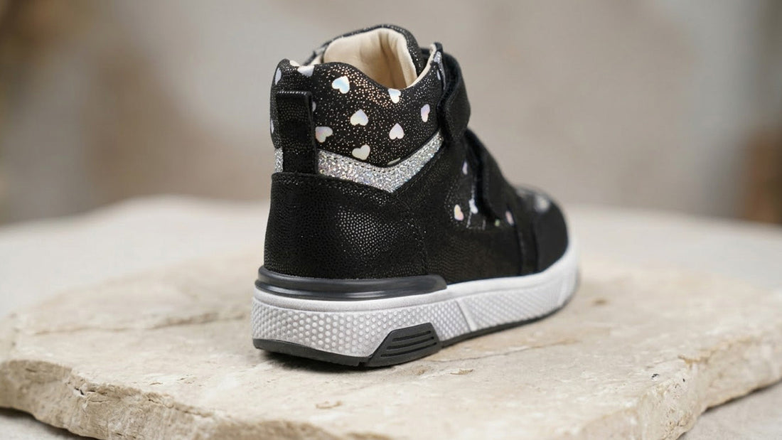 Rear perspective photograph of a children's orthopaedic shoe on a wooden floor demonstrating the flared, wide-base rubber outsole design.