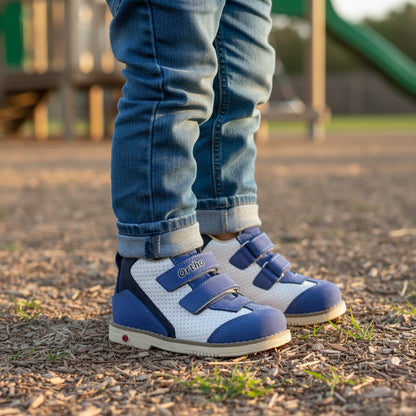 Child wearing blue jeans and Ortho Shoes orthopedic boots with white leather and blue suede accents, triple Velcro straps, arch and ankle support, and durable non-slip sole, standing on playground wood chips with slide and play structure in background.