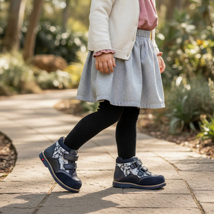 Young girl walking on a sunlit garden path in gray skirt and black tights, wearing silver black Ortho Shoes orthopedic boots with black and white floral pattern, Velcro straps, cushioned sole, Thomas heels, arch and ankle support.