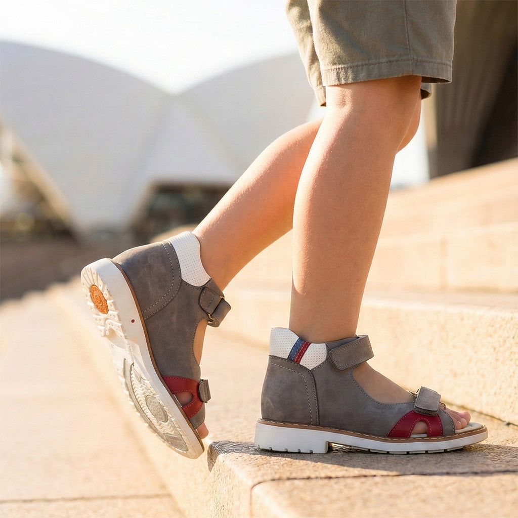 Boys’ orthopedic sandals in gray and red with mesh panels, Velcro straps, arch and ankle support, and Thomas heels, shown during outdoor play.