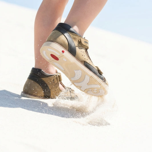 Child wearing grey and black orthopedic shoes with thick white soles, arch and ankle support, Thomas heels, kicking up sand while walking on a bright sandy surface.