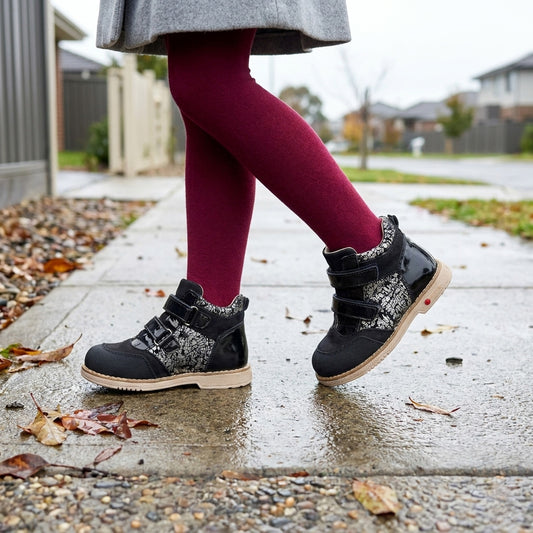 Girls’ black and silver orthopedic boots with Velcro straps, sturdy beige sole, arch and ankle support, Thomas heels and weather‑ready design, pictured on a rainy sidewalk.