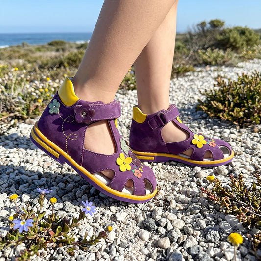 Children’s purple orthopedic sandals with flower details, yellow sole, and supportive structure, pictured on a rocky path near the coast.