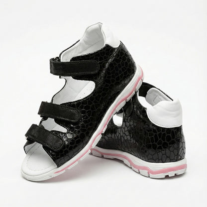 Children’s black orthopedic sandals with reptile-textured finish, white interior, and pink-accented sole.