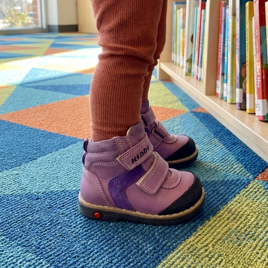 Purple children’s orthopedic boots with arch and ankle support, Thomas heels, dual Velcro straps, and dark toe caps, shown in a library reading area.
