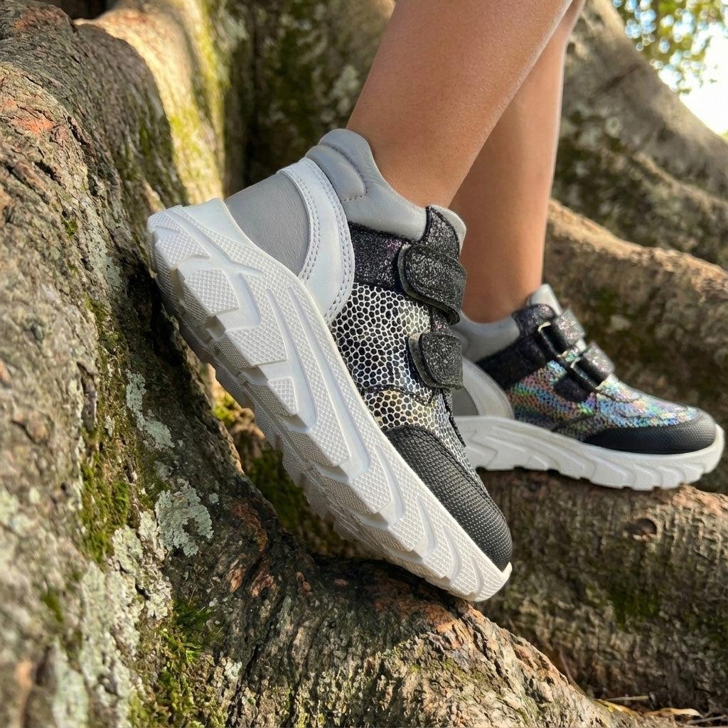 Child wearing silver-black orthopaedic mid-top sneakers with Velcro straps, arch and ankle support, and chunky white sole, standing on tree roots.