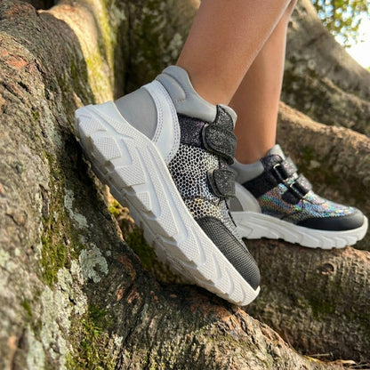 Child wearing silver-black orthopaedic mid-top sneakers with Velcro straps, arch and ankle support, and chunky white sole, standing on tree roots.