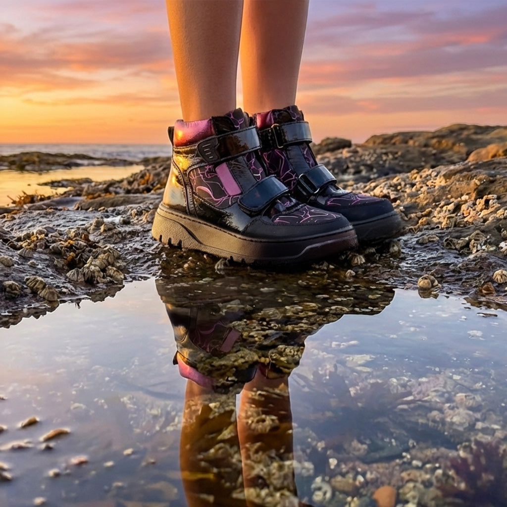 Child standing on rocky shoreline at sunset wearing black and purple high-top shoes with Velcro straps, arch and ankle support by Ortho Shoes.
