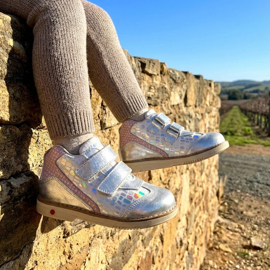 Child sitting on a stone wall wearing iridescent silver orthopedic boots with Velcro straps and supportive design, overlooking a vineyard.