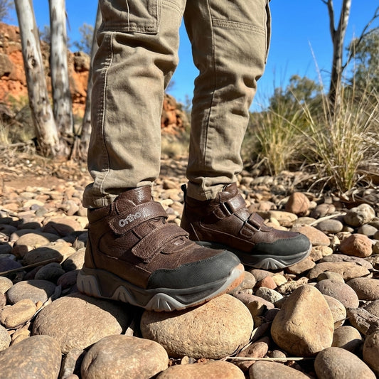 Child wearing brown orthopedic boots with Velcro straps, arch and ankle support, standing on rocky ground in a dry forest.