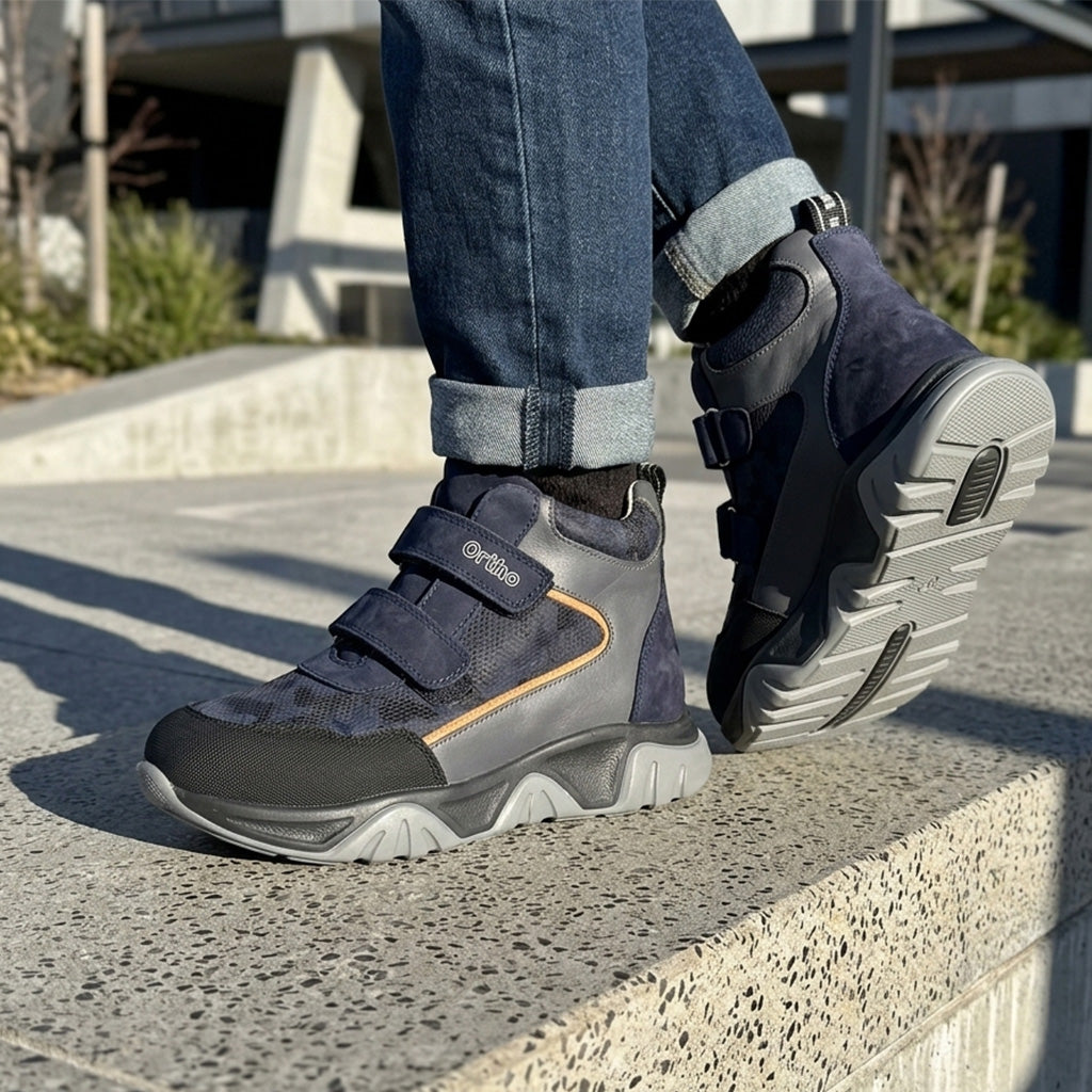 Children orthopedic boots in dark blue and grey with Velcro closure, cushioned textured sole, and supportive design, shown outdoors in an urban setting by Ortho Shoes.
