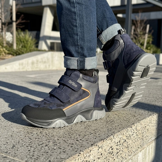 Children orthopedic boots in dark blue and grey with Velcro closure, cushioned textured sole, and supportive design, shown outdoors in an urban setting by Ortho Shoes.