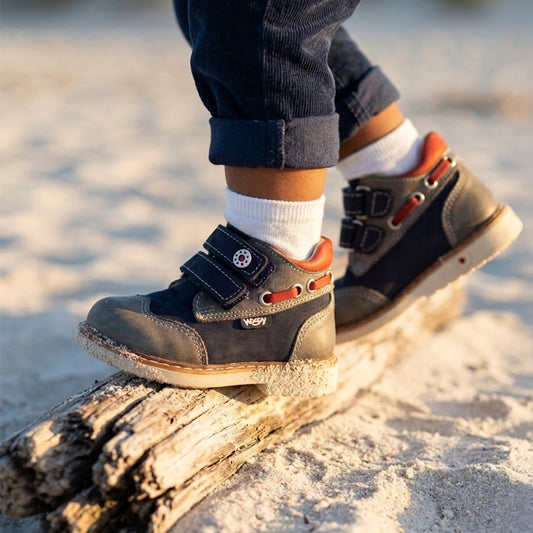 Child standing on a driftwood log wearing navy and grey orthopedic boots with Velcro straps, arch and ankles support, and Thomas heels by Woopy.