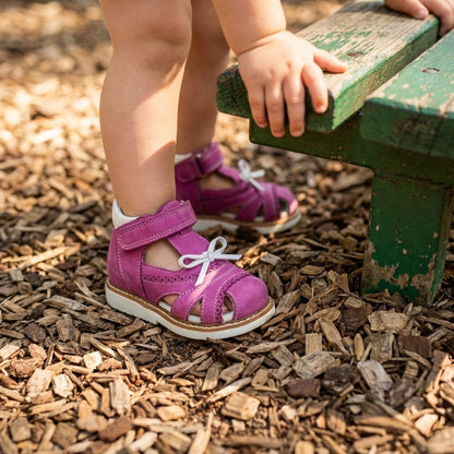 Toddler girl wearing bright purple orthopedic sandals with decorative laces and Velcro straps, arch and ankle support standing on wood chips near a green bench.