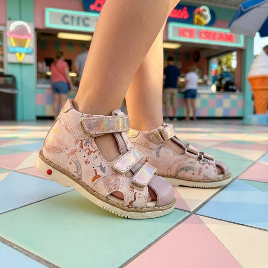Child wearing pink orthopedic sandals with iridescent straps, arch and ankle support. Thomas heels and underwater‑animal print, standing on a colourful tiled floor in an ice‑cream shop.