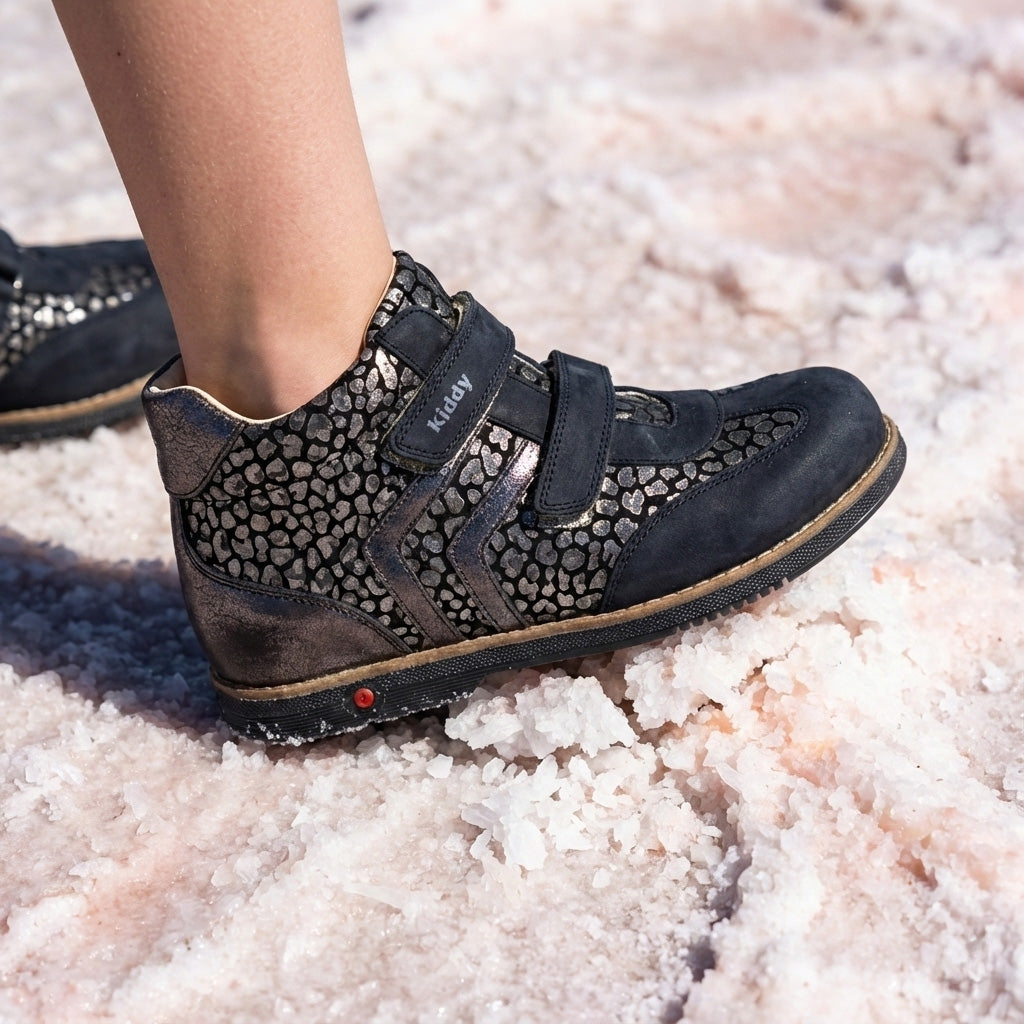 Child’s foot in dark orthopedic boot with Velcro straps and metallic pebble texture, arch and ankle support, and Thomas heel standing on coarse salt.