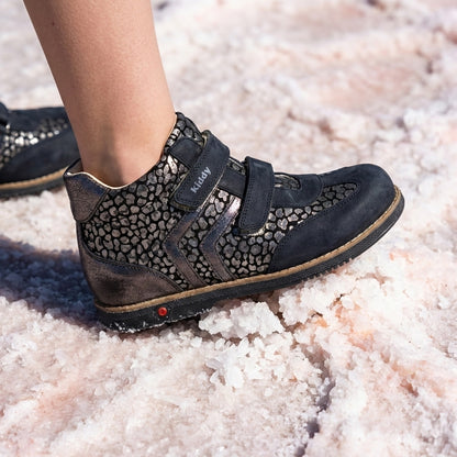 Child’s foot in dark orthopedic boot with Velcro straps and metallic pebble texture, arch and ankle support, and Thomas heel standing on coarse salt.