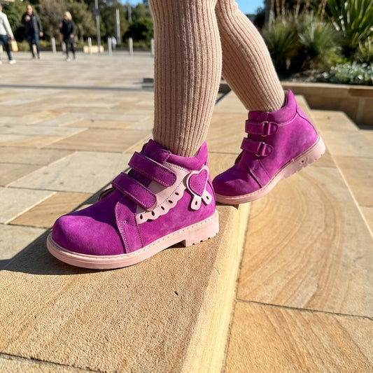 Child standing on a stone step wearing bright magenta orthopaedic boots with pink elements, heart detail, Velcro straps, arch and ankle support, Thomas heels by Woopy.
