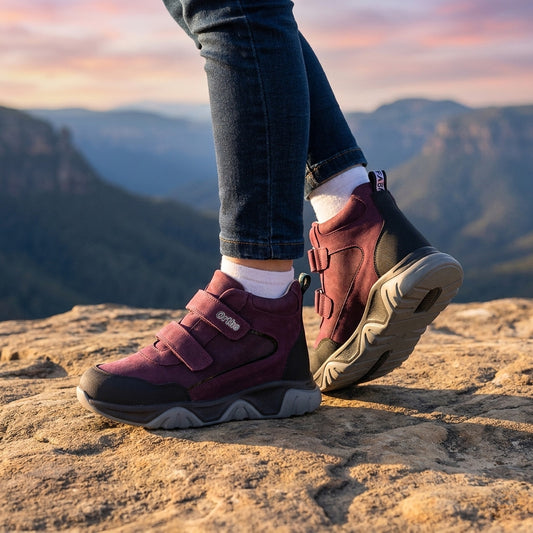 Dark purple kids orthopedic boots with sturdy sole, Velcro straps, and reinforced support, shown on rocky terrain at sunset.
