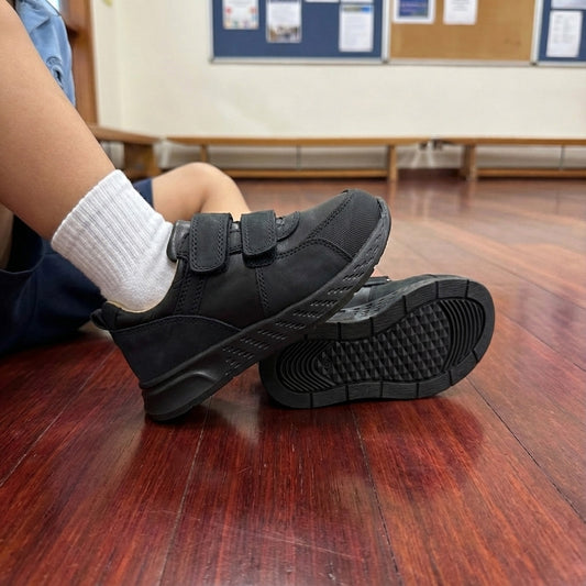 Child seated indoors wearing low-top black Velcro-strap orthopaedic sneakers with arch and ankle support and white socks on polished wooden flooring.